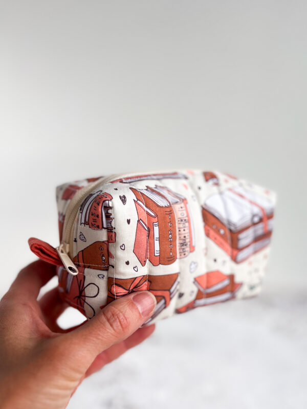 Pikowana kosmetyczka handmade w książkowy wzór z ilustracjami książek, zapinana na zamek, na jasnym tle w klimacie cozy.