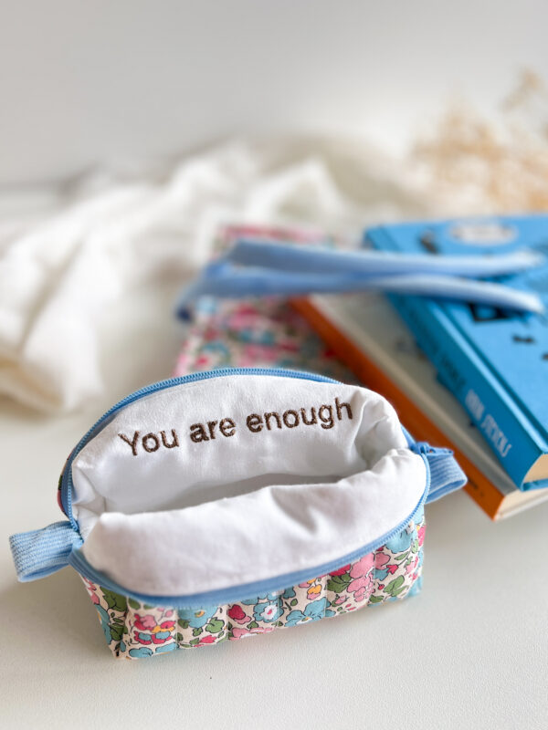 Mała kosmetyczka w kwiatowy wzór z widocznym haftem „You are enough” na jasnej podszewce, na tle książek.